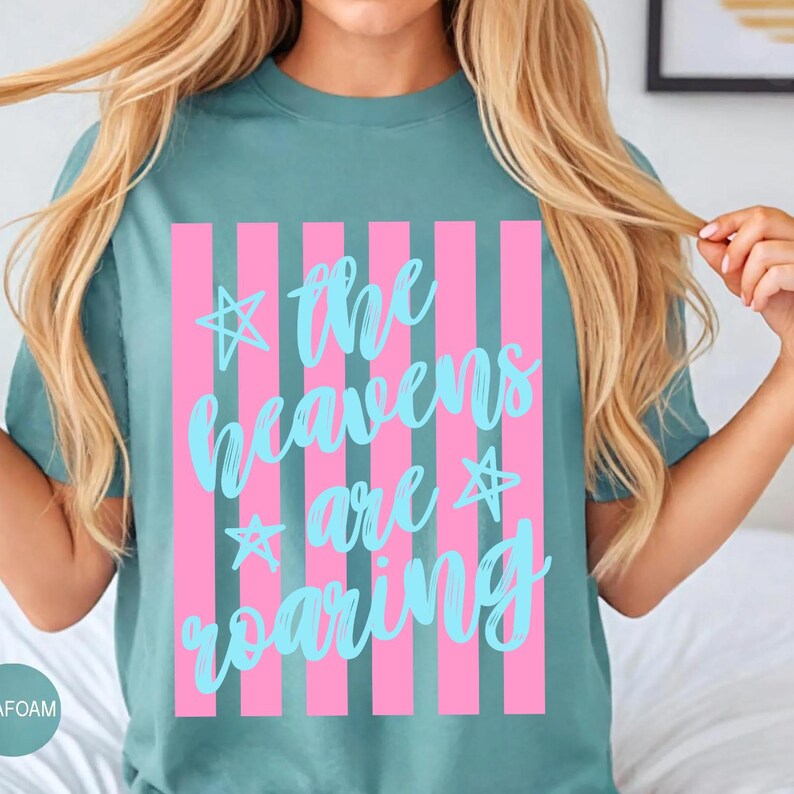 May include: Teal-colored t-shirt with a pink striped design. The shirt features the text "the heavens are roaring" in light blue script, with star accents. A casual, comfortable top.