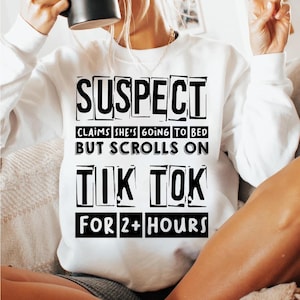 Może przedstawiać: Biała bluza z czarnym nadrukiem, który brzmi "SUSPECT CLAIMS SHE'S GOING TO BED BUT SCROLLS ON TIK TOK FOR 2+ HOURS".