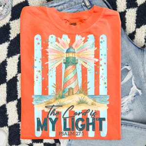 Pu&ograve; includere: T-shirt arancione con un design a faro colorato e la scritta "The Lord is My Light" e "Psalm 27:1". Il faro ha un motivo a strisce ed &egrave; ambientato su uno sfondo a strisce blu e arancioni. La maglietta &egrave; piegata.