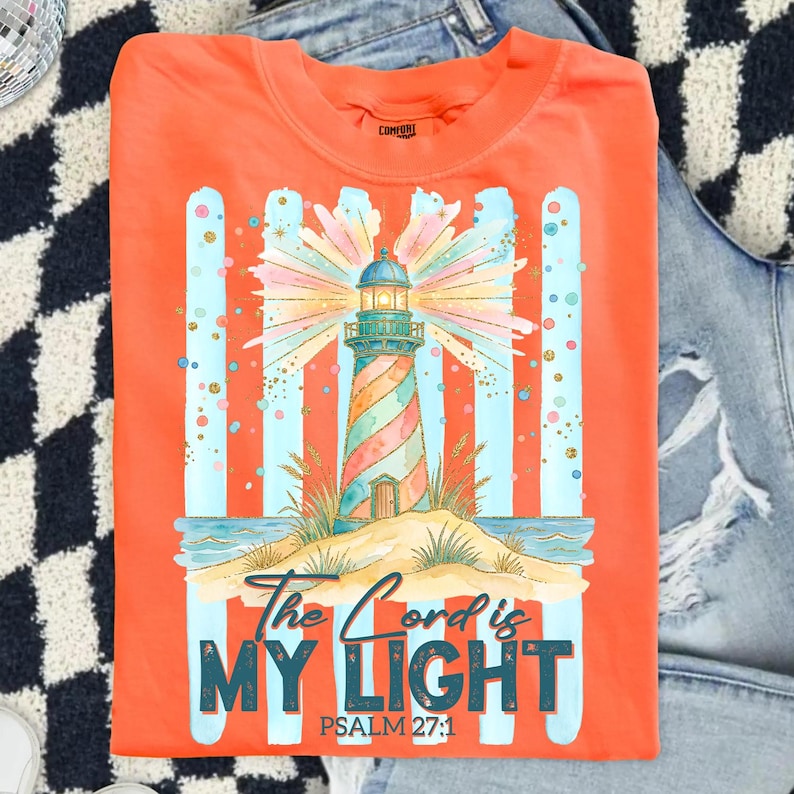 Pu&ograve; includere: T-shirt arancione con un design a faro colorato e la scritta "The Lord is My Light" e "Psalm 27:1". Il faro ha un motivo a strisce ed &egrave; ambientato su uno sfondo a strisce blu e arancioni. La maglietta &egrave; piegata.