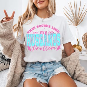 May include: White t-shirt with the text "THAT SOUNDS LIKE my HUSBANDS Problem" in blue and pink. The shirt is worn with denim shorts and a beige cardigan. The person is giving a peace sign.