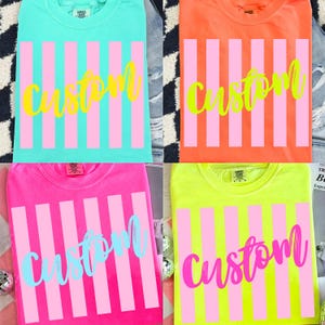 K&ouml;nnte beinhalten: Vier farbenfrohe T-Shirts: T&uuml;rkis, Orange, Pink und Neongelb. Jedes Shirt hat vertikale rosa Streifen und das Wort "custom" in einer Kontrastfarbe. Die Shirts sind gefaltet und auf einer gemusterten Oberfl&auml;che ausgestellt.