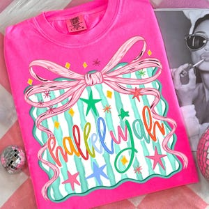 May include: Bright pink Comfort Colors t-shirt with a colorful graphic. The graphic features a pink bow, stars, and the word "hallelujah" in a rainbow of colors. The shirt is a crew neck and has a relaxed fit.