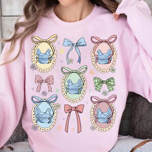 May include: A light pink sweatshirt featuring a repeating pattern of decorative bows and oval frames. Each frame contains a blue chicken illustration. The bows are in various colors, including pink, blue, and green.