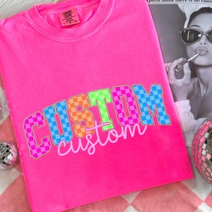 May include: Bright pink t-shirt with the word "CUSTOM" in a checkered pattern of pink, orange, green, and blue. The word "custom" is written below in a cursive font. The shirt is a Comfort Colors brand.