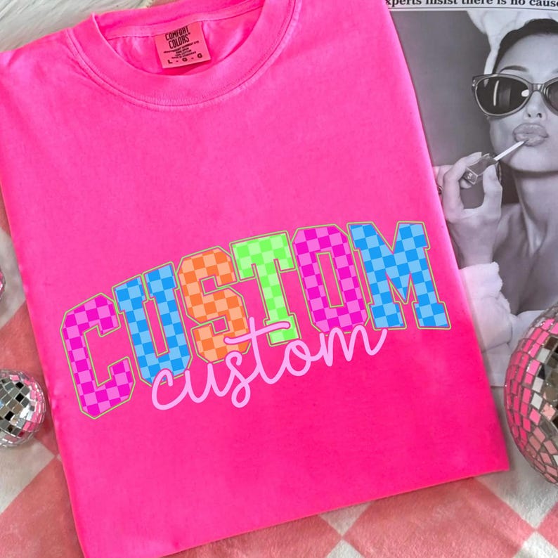 May include: Bright pink t-shirt with the word "CUSTOM" in a checkered pattern of pink, orange, green, and blue. The word "custom" is written below in a cursive font. The shirt is a Comfort Colors brand.