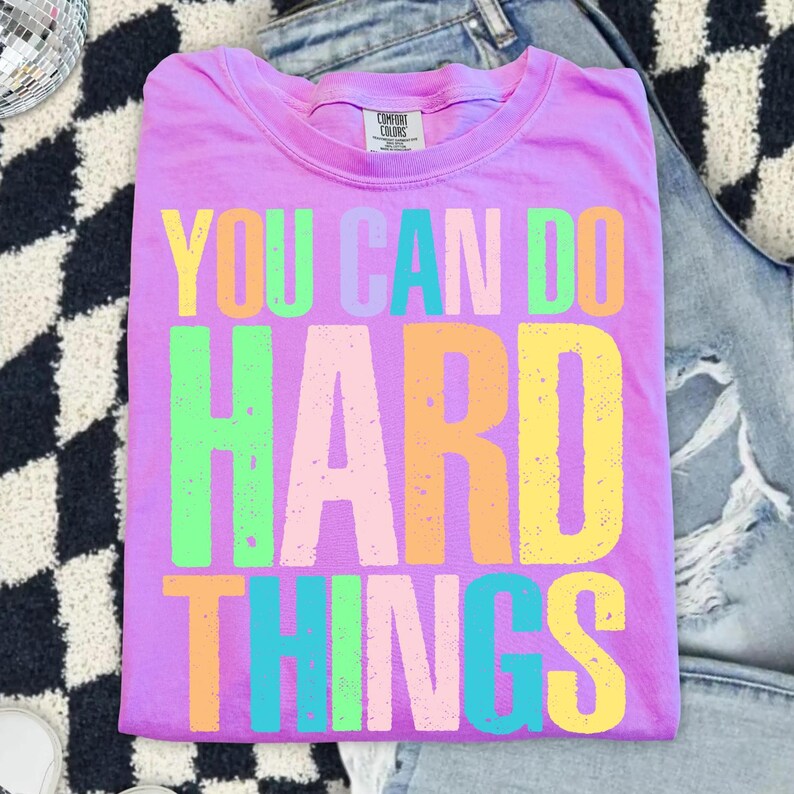 May include: A lilac-colored t-shirt with the phrase "YOU CAN DO HARD THINGS" printed in large, colorful letters. The text is in yellow, green, pink, orange, and blue. The shirt is folded and placed on a black and white patterned surface.