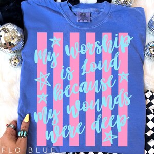 May include: Flo blue t-shirt with pink vertical stripes and the text "My worship is loud because my wounds were deep" in light blue. The shirt has a crew neck and short sleeves. The shirt is made of a soft, comfortable material.