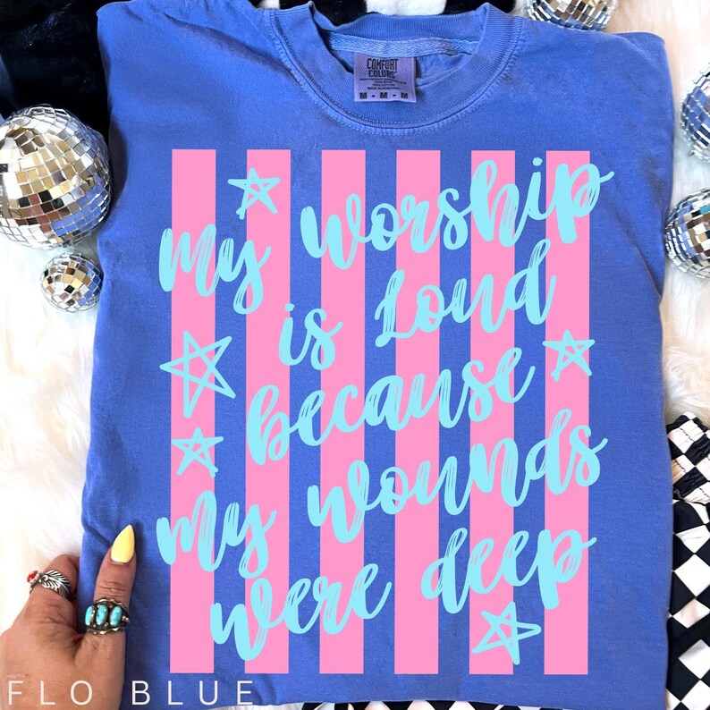 May include: Flo blue t-shirt with pink vertical stripes and the text "My worship is loud because my wounds were deep" in light blue. The shirt has a crew neck and short sleeves. The shirt is made of a soft, comfortable material.