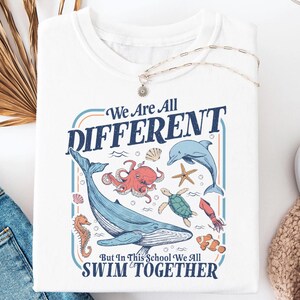May include: White t-shirt with a colorful graphic of various sea creatures, including a whale, octopus, and dolphin. The text reads "We Are All DIFFERENT But In This School We All SWIM TOGETHER."