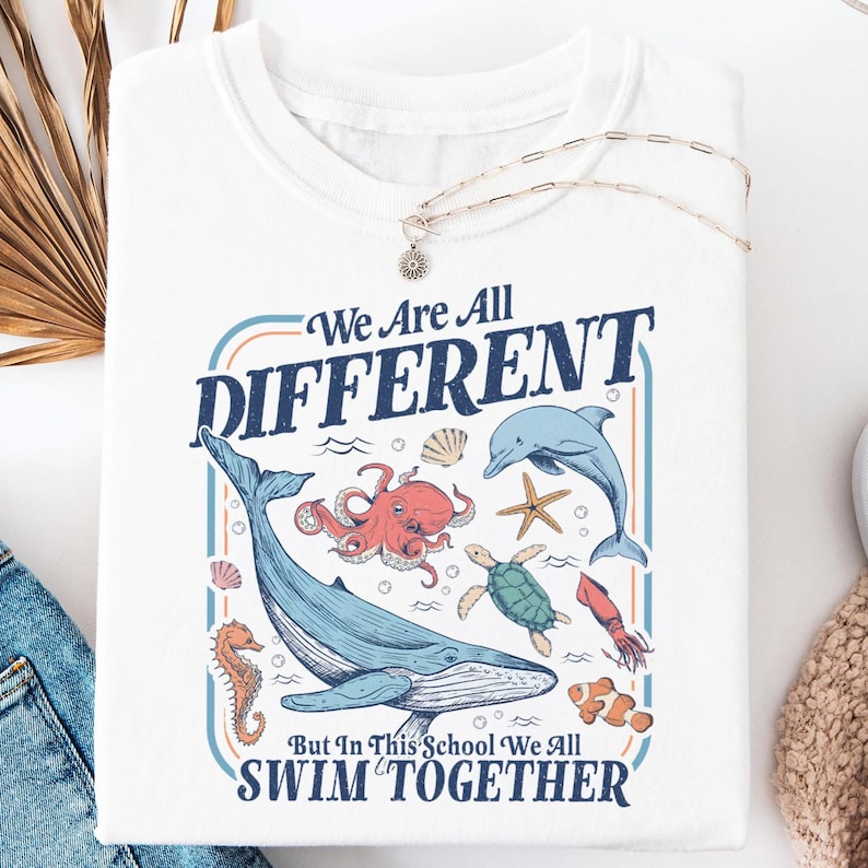 May include: White t-shirt with a colorful graphic of various sea creatures, including a whale, octopus, and dolphin. The text reads "We Are All DIFFERENT But In This School We All SWIM TOGETHER."