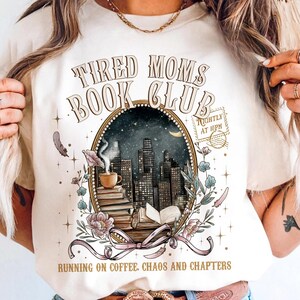 May include: Cream-colored t-shirt with the text "Tired Moms Book Club" in a vintage style. The design features a cityscape inside an oval frame, a coffee cup, books, and floral accents. The text "Running on coffee, chaos and chapters" is at the bottom.