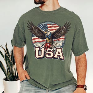 May include: Olive green t-shirt featuring a graphic of an eagle with outstretched wings, wearing medals, and perched atop a red and white object. The background includes a U.S. flag and the word "USA" in bold letters.