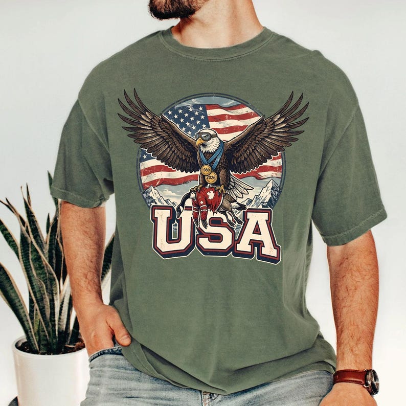 May include: Olive green t-shirt featuring a graphic of an eagle with outstretched wings, wearing medals, and perched atop a red and white object. The background includes a U.S. flag and the word "USA" in bold letters.
