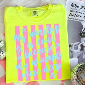 May include: A neon yellow t-shirt with the text "My worship is loud because my wounds were deep" in light blue script and pink vertical stripes. The shirt is a Comfort Colors brand, size L-G.