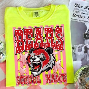 May include: A neon yellow t-shirt with a graphic design featuring the word "BEARS" in red letters filled with white stars. Below the word "BEARS" is a bear wearing star-shaped sunglasses. The words "SCHOOL NAME" are at the bottom.