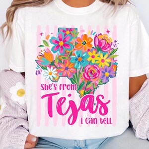 May include: White t-shirt featuring a colorful floral design in the shape of Texas. The design includes a variety of flowers in shades of pink, orange, blue, and yellow. The text "she's from Tejas I can tell" is written in pink.