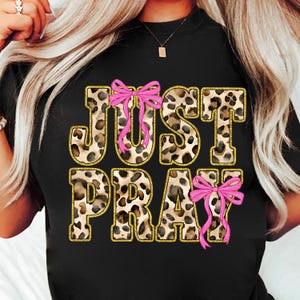 May include: Black t-shirt featuring the words "JUST PRAY" in a leopard print design with gold glitter outlines. Pink bows are placed above the "U" and below the "Y" in the text.