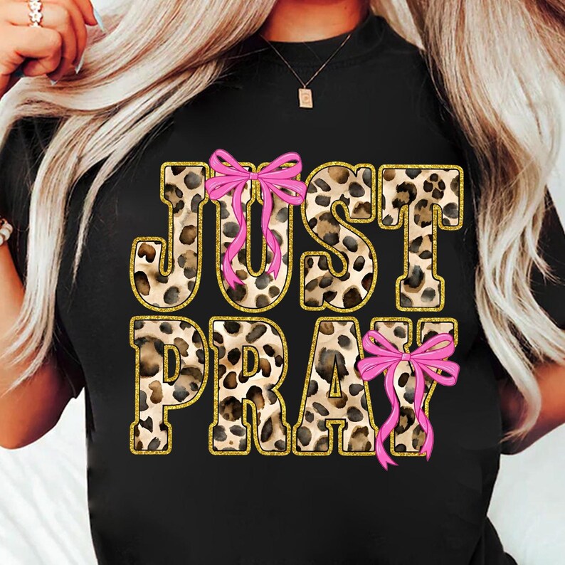 May include: Black t-shirt featuring the words "JUST PRAY" in a leopard print design with gold glitter outlines. Pink bows are placed above the "U" and below the "Y" in the text.