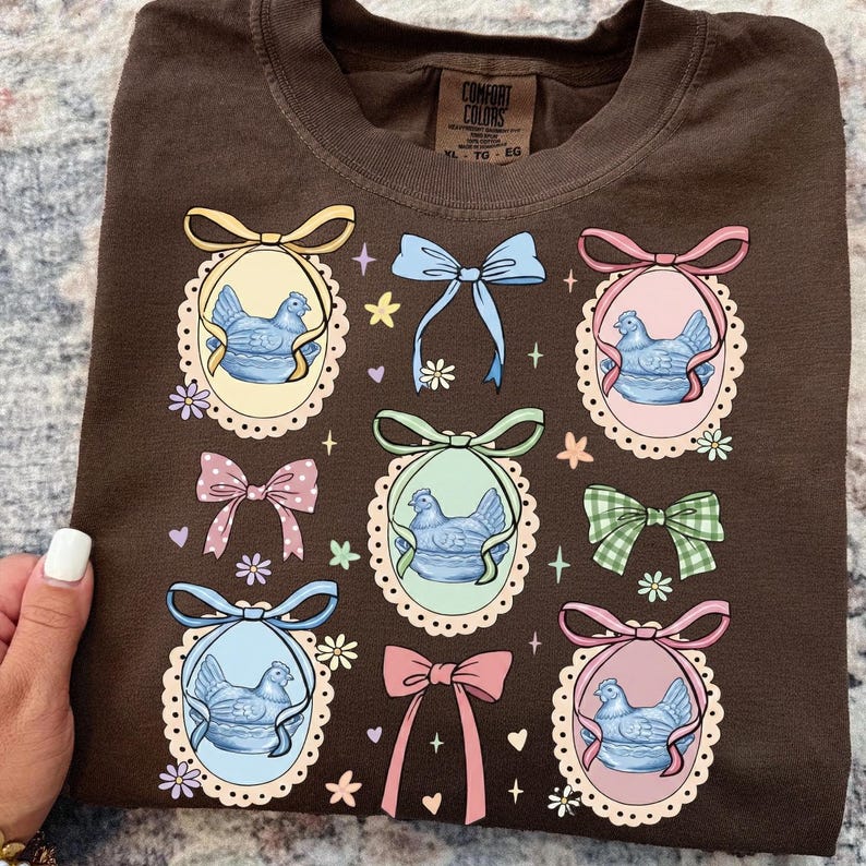 May include: A brown Comfort Colors t-shirt featuring a repeating pattern of blue hen illustrations within ornate frames, surrounded by colorful bows and floral accents. The shirt has a relaxed fit and a vintage aesthetic.