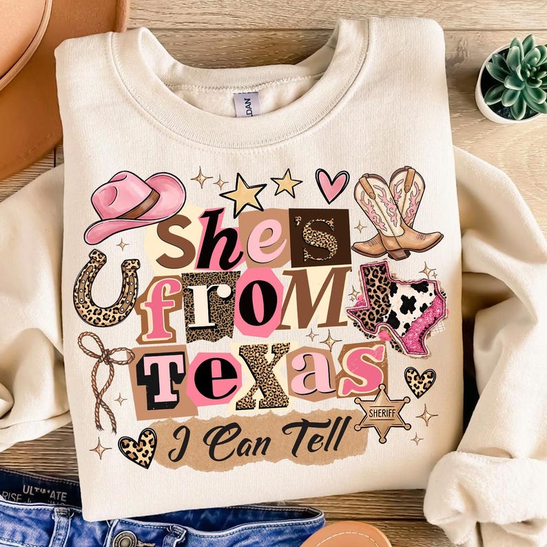 May include: Cream-colored sweatshirt with a graphic design featuring a pink cowboy hat, boots, and the text "She's From Texas I Can Tell." The design includes stars, hearts, and a horseshoe.