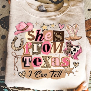 May include: A light beige sweatshirt with a graphic design that reads "She's From Texas I Can Tell." The design includes a pink cowboy hat, boots, a horseshoe, stars, hearts, and the outline of Texas. The text and graphics are in shades of pink, brown, and leopard print.
