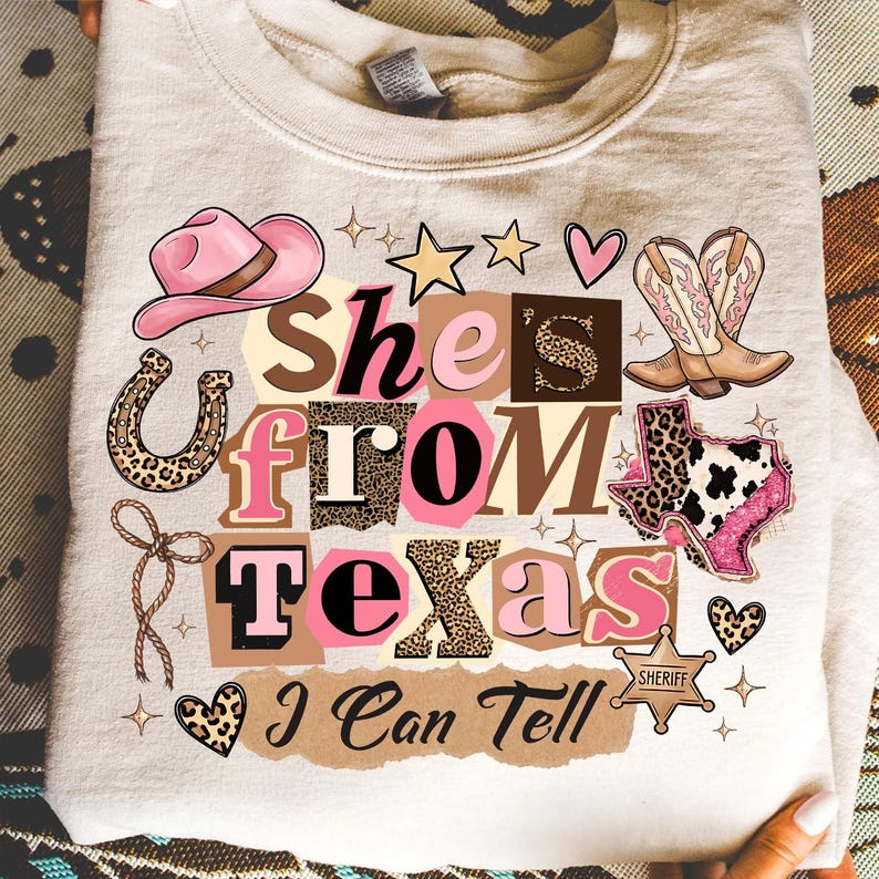 May include: A light beige sweatshirt with a graphic design that reads "She's From Texas I Can Tell." The design includes a pink cowboy hat, boots, a horseshoe, stars, hearts, and the outline of Texas. The text and graphics are in shades of pink, brown, and leopard print.