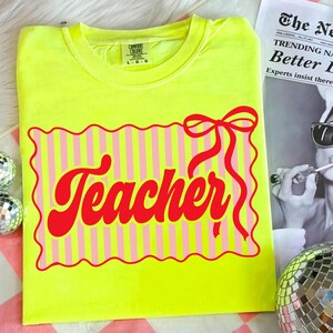 May include: A neon yellow t-shirt with a red and pink graphic that says "Teacher". The graphic is a rectangle with pink and white stripes and a red bow. The shirt is a Comfort Colors brand.