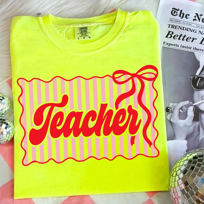 May include: A neon yellow t-shirt with a red and pink graphic that says "Teacher". The graphic is a rectangle with pink and white stripes and a red bow. The shirt is a Comfort Colors brand.
