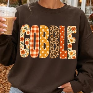 May include: A dark brown long-sleeve sweatshirt featuring the word GOBBLE printed in large letters across the chest. Each letter has a distinct pattern: floral, orange and white gingham, yellow polka dot, leopard print, and orange polka dot.