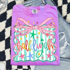 May include: A lilac Comfort Colors t-shirt featuring a colorful graphic. The graphic includes a pink bow, teal and white striped background, and the word "Hallelujah" in rainbow colors, with stars and diamond accents.