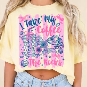 May include: Yellow t-shirt with a graphic that says "I take my coffee on the rocks." The graphic includes a drink, hat, sunglasses, seashells, and tropical flowers in pink and blue.