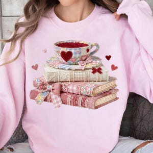 May include: Pink sweatshirt featuring a stack of books topped with a teacup. The teacup and books are decorated with floral and heart patterns. A red heart is on the teacup. Small heart accents are scattered around the design.