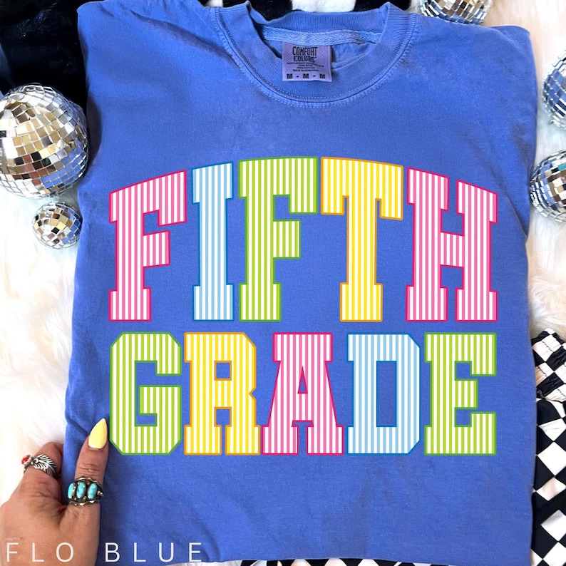 May include: A periwinkle blue t-shirt featuring the text FIFTH GRADE in a bold, collegiate-style font. Each letter is filled with a vertical white pinstripe pattern and colored in alternating shades of pink, light blue, lime green, and yellow.