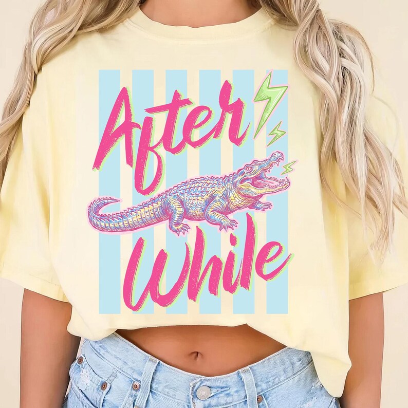 Pode incluir: Uma camiseta amarelo claro com as palavras "After While" em escrita rosa, apresentando uma ilustra&ccedil;&atilde;o colorida de jacar&eacute; em um fundo listrado azul. Raios verdes acentuam o design.