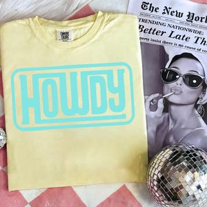 May include: A light yellow t-shirt with the word "HOWDY" in a light blue, retro-style font. The shirt is made of a soft material and has a crew neck. The shirt is displayed on a pink and white checkered surface.