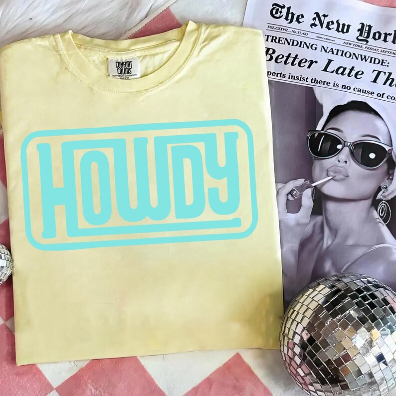 May include: A light yellow t-shirt with the word "HOWDY" in a light blue, retro-style font. The shirt is made of a soft material and has a crew neck. The shirt is displayed on a pink and white checkered surface.