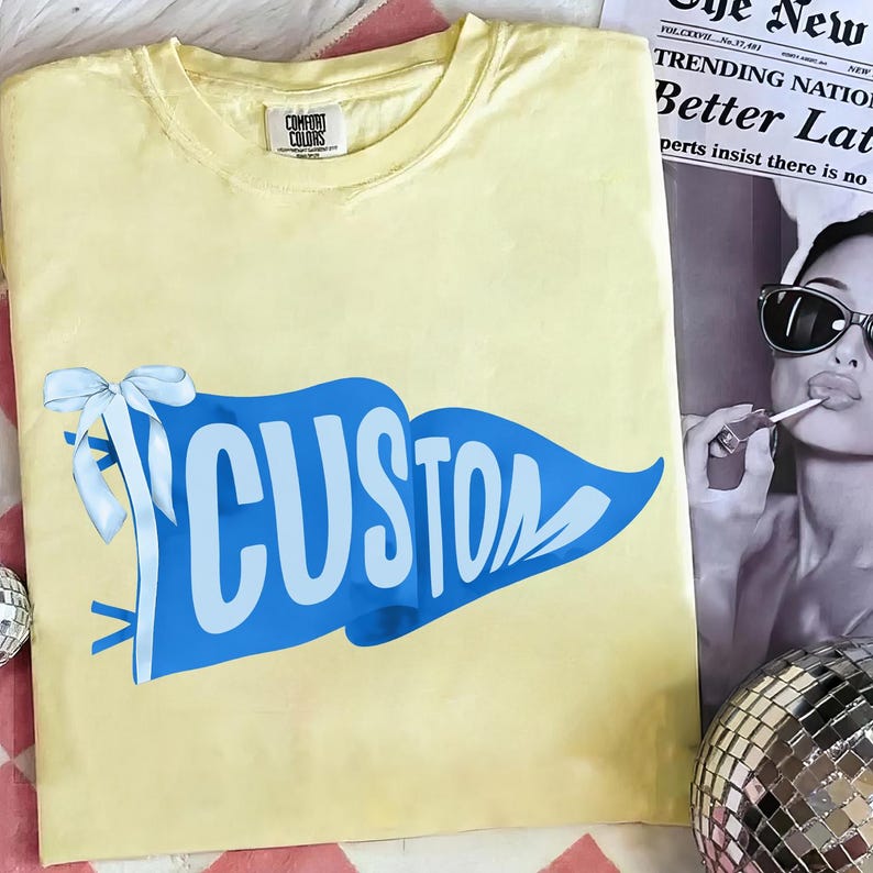 May include: A light yellow t-shirt with a blue pennant design that says "CUSTOM" in white letters. The pennant has a white bow on the left side. The shirt is made by Comfort Colors.