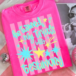 May include: Bright pink t-shirt with the text "He turned my trials into testimony" in blue and yellow. The design includes vertical pink stripes and star graphics. The shirt is a Comfort Colors brand.