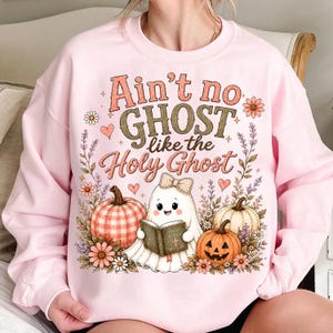 May include: A light pink crewneck sweatshirt featuring a graphic print with the text "Ain't no GHOST like the Holy Ghost". The design includes a cartoon ghost wearing a bow, holding a Bible, surrounded by pumpkins, flowers, and small hearts.
