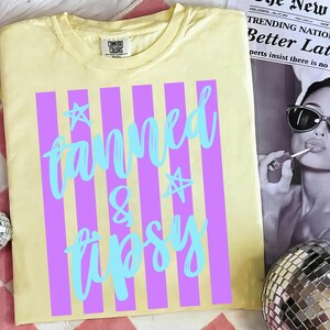Może przedstawiać: Jasnoż&oacute;łta koszulka z pionowymi fioletowymi paskami i napisem "fanned & tipsy" w jasnoniebieskim skrypcie. Koszulka posiada r&oacute;wnież gwiazdki. Koszulka jest marki Comfort Colors.