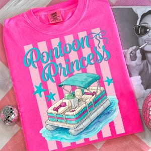 May include: A hot pink t-shirt with the words "Pontoon Princess" in turquoise script. The shirt features a graphic of a pontoon boat with a teal canopy and blue stars. The shirt is a Comfort Colors brand.