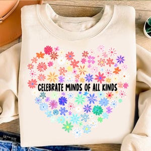 May include: A beige sweatshirt with a heart-shaped design made of colorful flowers. The text "Celebrate Minds of All Kinds" is printed below the heart.