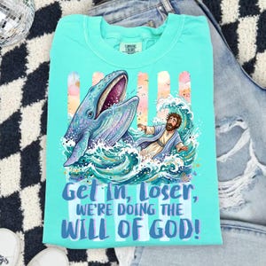 May include: A turquoise Comfort Colors t-shirt featuring a graphic of a whale, a man in a robe, and ocean waves. The text "Get In, Loser, We're Doing The Will Of God!" is printed in blue. The shirt is folded on a denim surface.