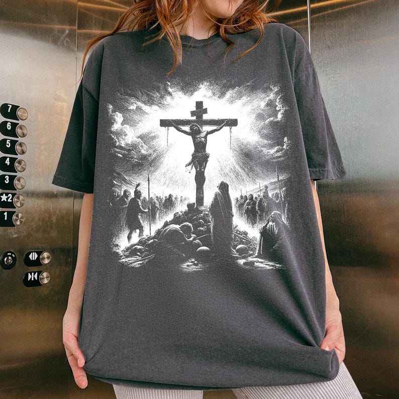 May include: Dark gray t-shirt featuring a detailed black and white illustration of the crucifixion of Jesus Christ. The graphic depicts Jesus on the cross, surrounded by figures and a dramatic sky. The shirt has a vintage, worn look.