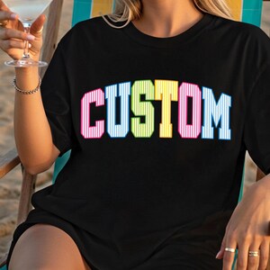 May include: Black t-shirt with the word "CUSTOM" in large, colorful, striped letters. The letters are outlined in white and have a retro, collegiate style. The shirt is being worn by a person.