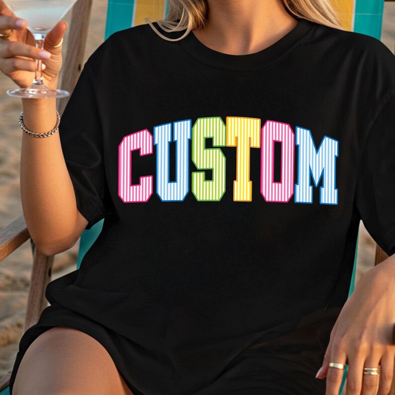 May include: Black t-shirt with the word "CUSTOM" in large, colorful, striped letters. The letters are outlined in white and have a retro, collegiate style. The shirt is being worn by a person.