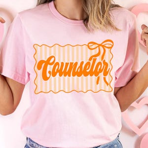 May include: A light pink t-shirt with the word "Counselor" in orange script. The text is inside a rectangular frame with a striped pattern and a bow on top. The shirt has short sleeves.