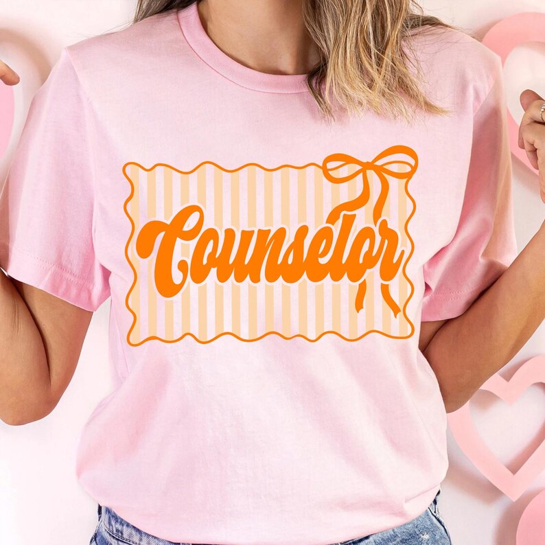 May include: A light pink t-shirt with the word "Counselor" in orange script. The text is inside a rectangular frame with a striped pattern and a bow on top. The shirt has short sleeves.