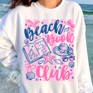 May include: White sweatshirt with a pink and blue graphic design. The design includes the words "Beach Book Club", seashells, a starfish, a hat, and sunglasses. The text "Members only dive into a good book" is also included.